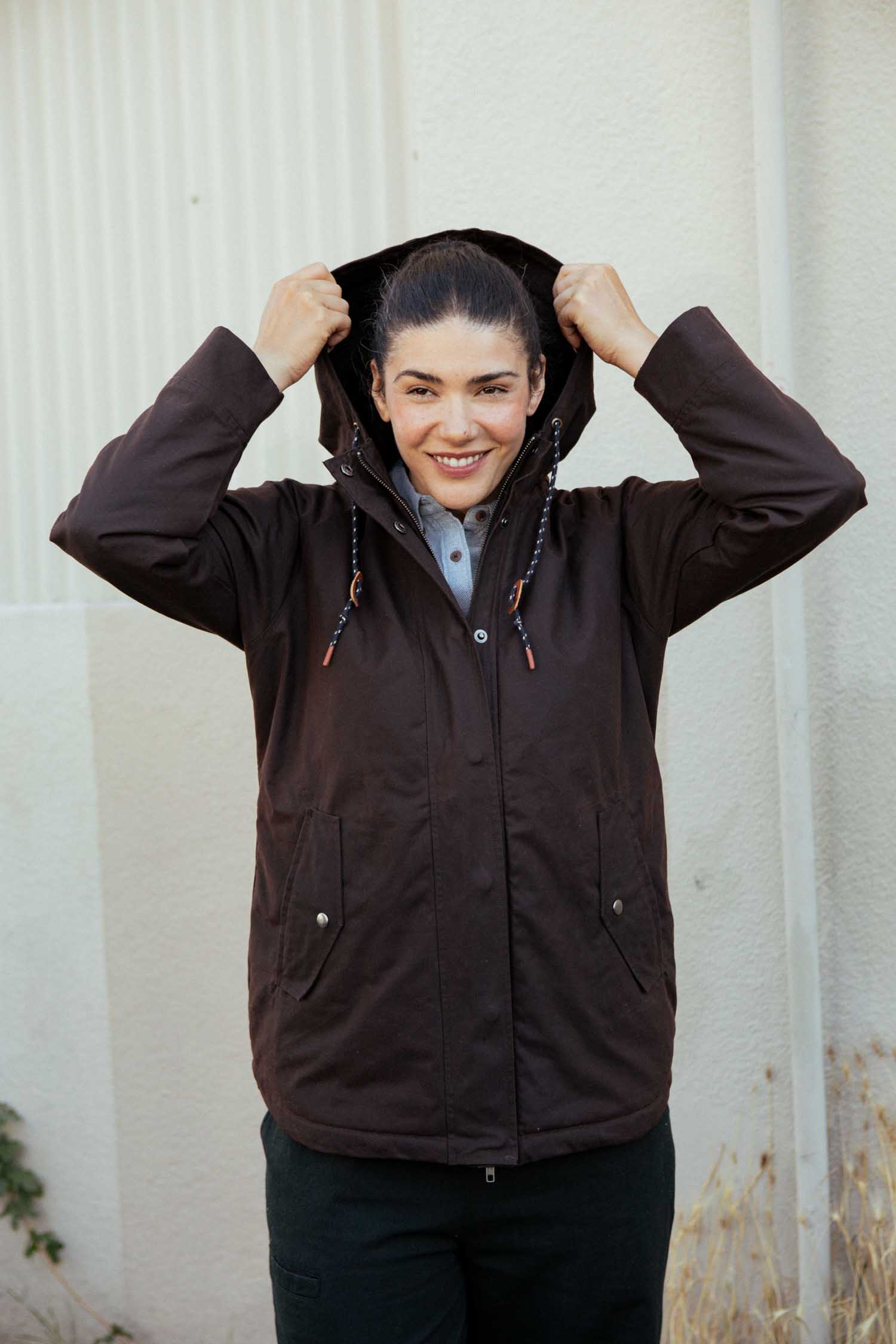 A person stands smiling outdoors, holding up the hood of the Bridge & Burn Sequoia Parka in Umber Waxed Canvas. They wear a light shirt underneath, with light walls and dry grass in the background.
