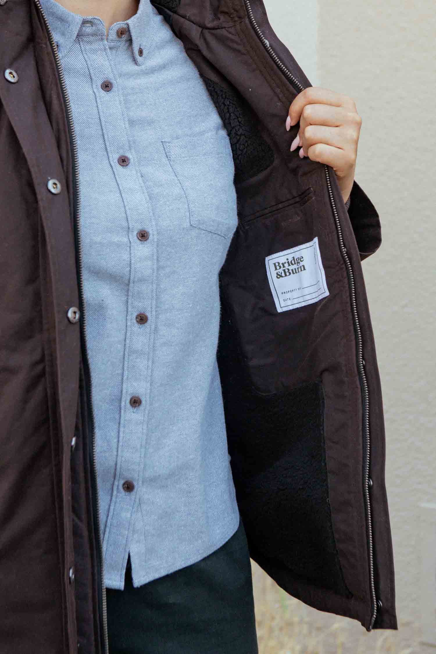 A person wearing a light blue shirt and the Bridge & Burn Sequoia Parka in Umber Waxed Canvas holds open the jacket to show the Bridge & Burn label sewn inside.