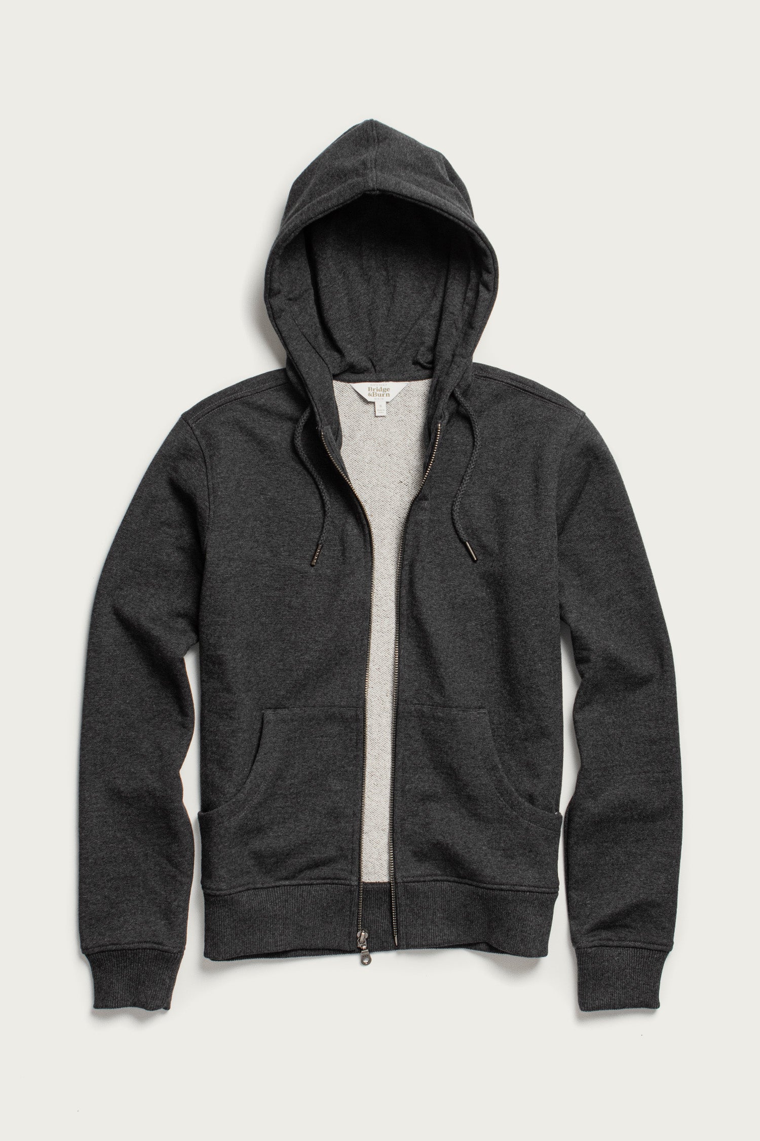 The Mason Hoodie in Graphite by Bridge & Burn is a dark gray French terry zip-up with front pockets, hood, drawstrings, and center zipper, shown flat on a light background.