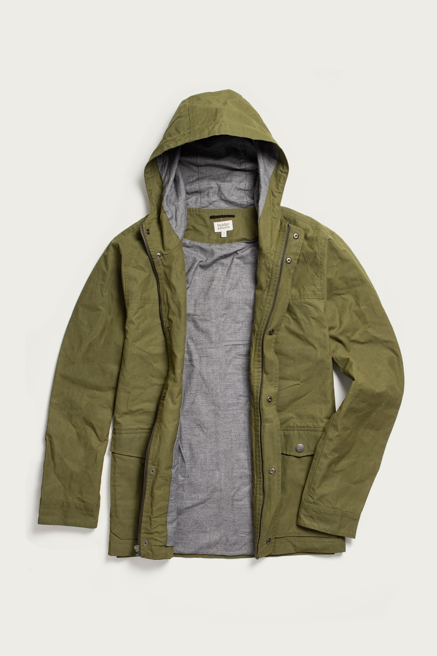 The Bridge & Burn Marshall Jacket in olive waxed canvas is a water-resistant, hooded jacket with a gray lining, snap buttons, and front pockets, shown unzipped and laid flat on a white background.