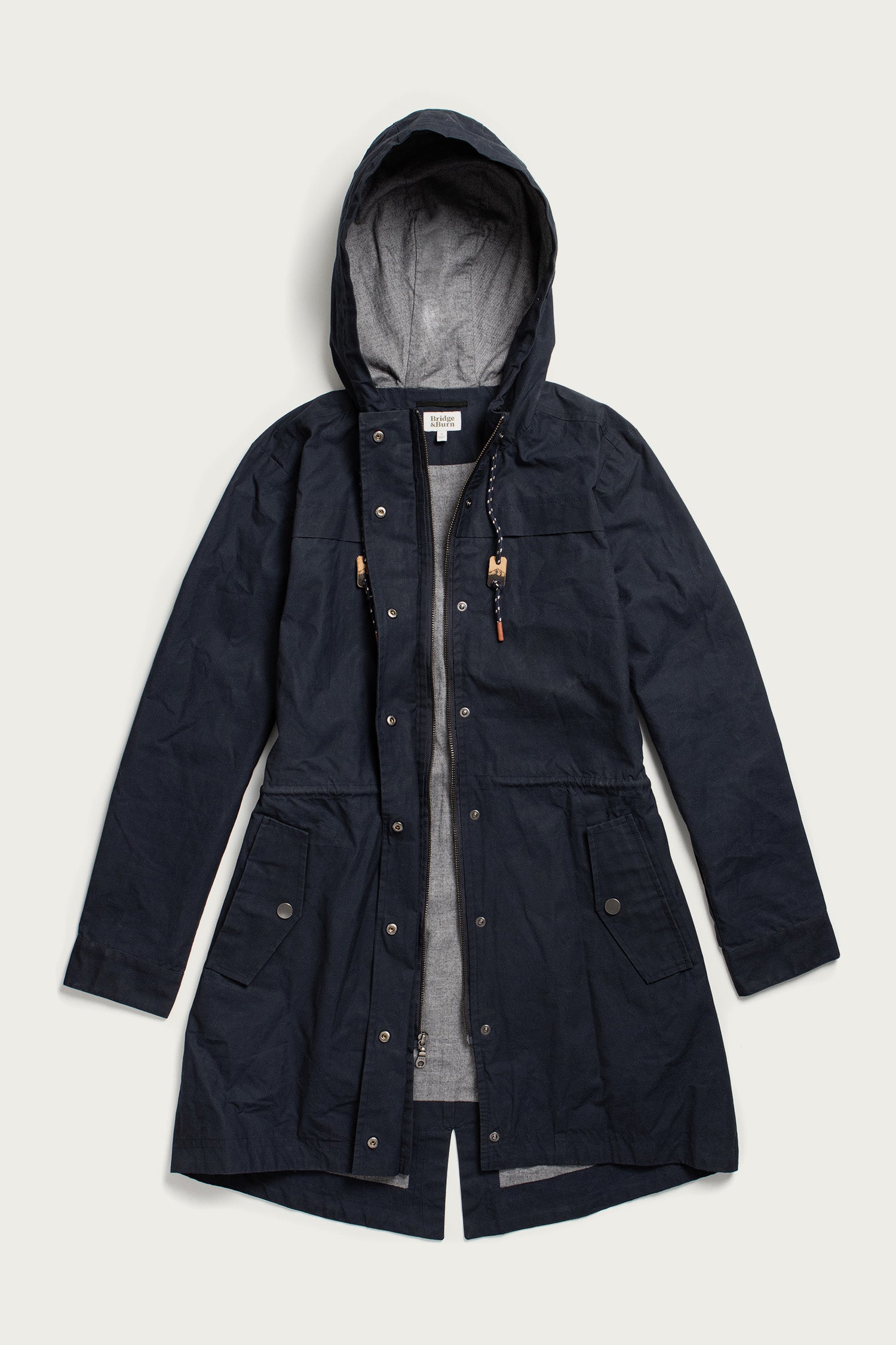 The Briar Jacket by Bridge & Burn is a navy waxed canvas parka with a hood, front zipper and snaps, two front flap pockets, adjustable waist and neck drawstrings, and a light gray lining—shown laid flat on a white background.