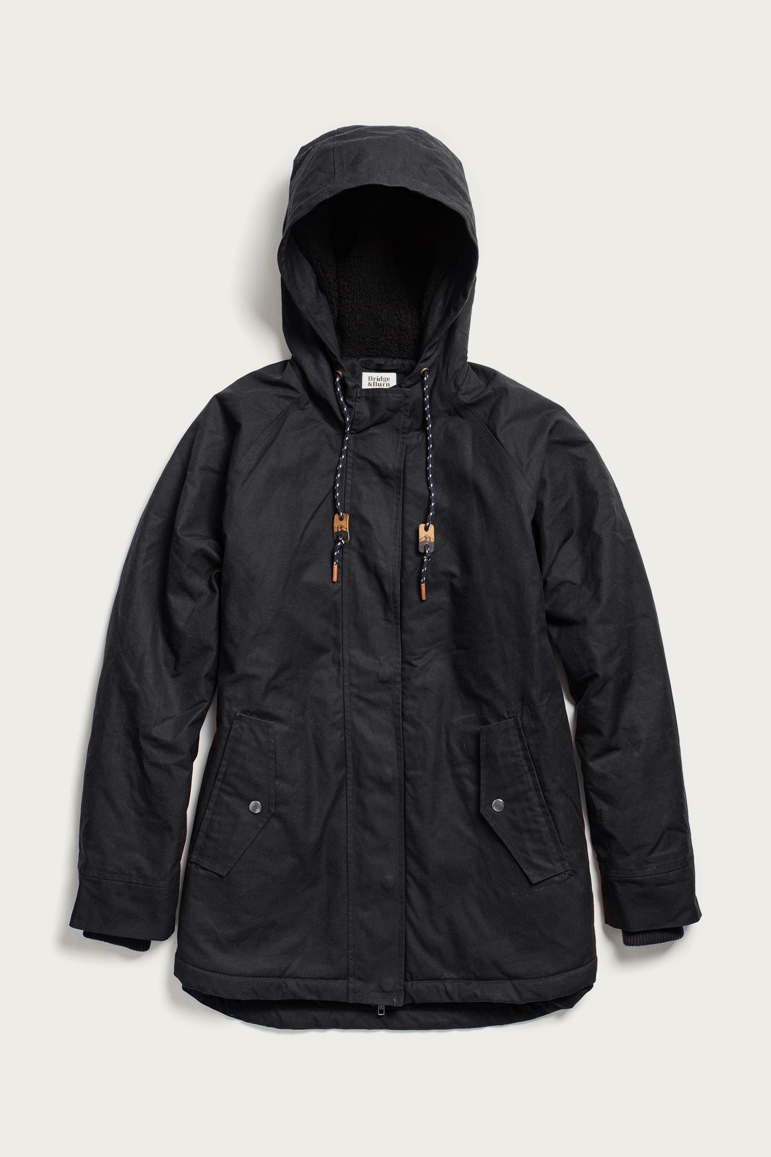 The Bridge & Burn Sequoia Parka in black waxed canvas features a hood with drawstrings, zip and button closure, two front flap pockets, ribbed cuffs, and soft fleece lining, shown laid flat against a light background.