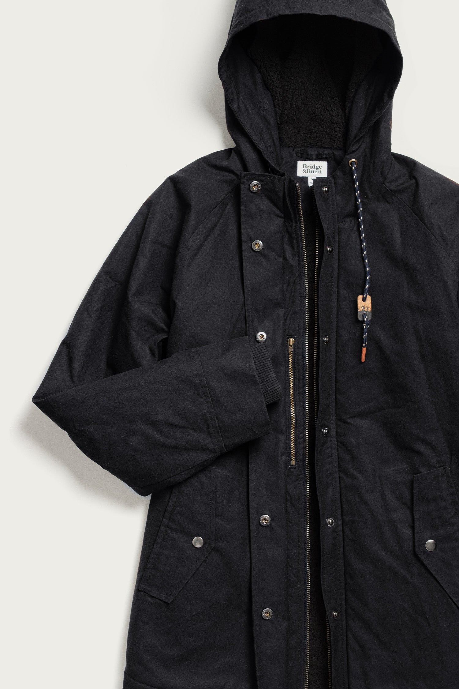 The Bridge & Burn Sequoia Parka in black waxed canvas features snap buttons, a zipper closure, side pockets, and a hood with a braided drawstring and wooden bead, shown on a light background.