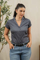 A woman with dark hair in a ponytail wears the Bridge & Burn Innes Shirt in Light Navy and blue jeans with a black belt, standing with her hands in her pockets next to a leafy plant against a neutral background.
