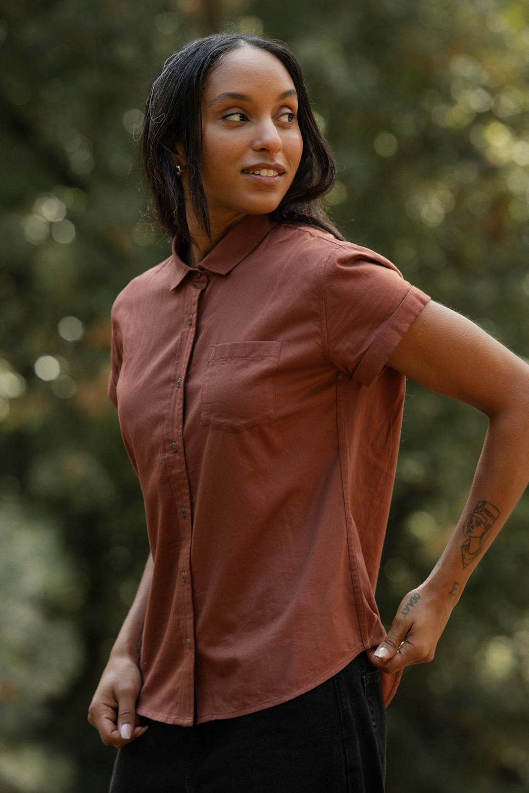 Wearing the Bridge & Burn Bea Shirt in Copper, a person stands outdoors smiling and looking to the side. The tailored, rust-colored short-sleeve button-down highlights their style; a forearm tattoo appears against blurred greenery.