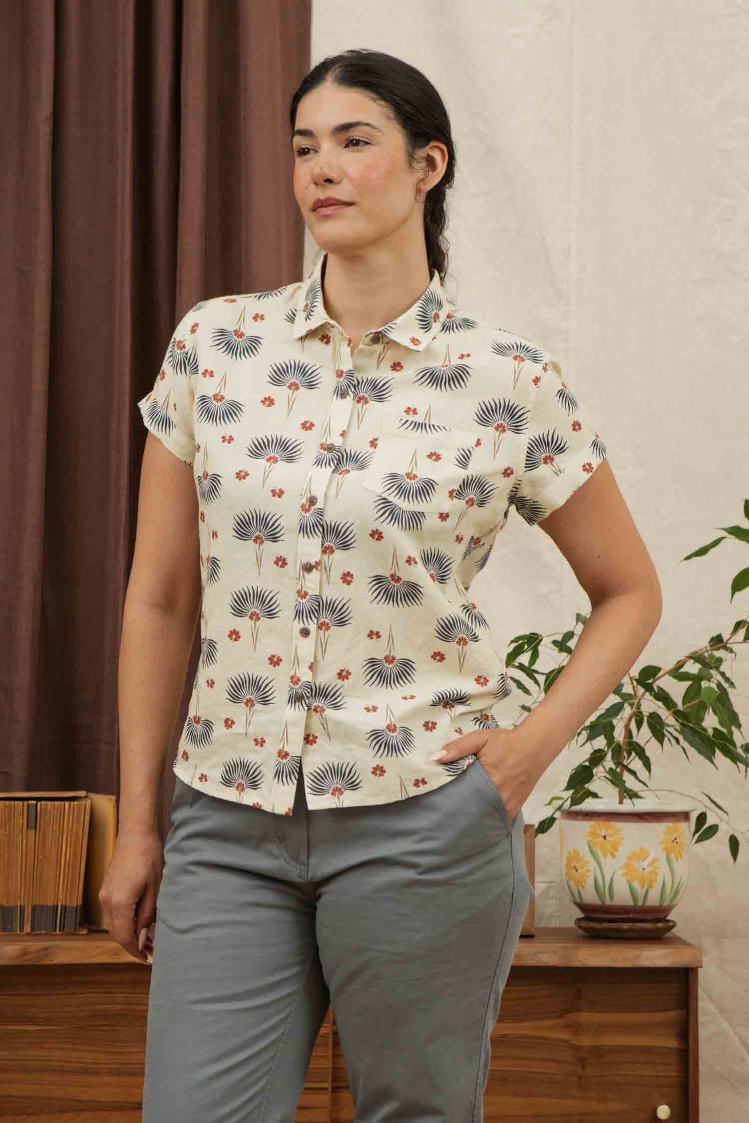 A woman with dark hair tied back wears the Bridge & Burn Bea Shirt in Aloha Floral, a short-sleeve button-down linen cotton shirt, and gray pants. She stands indoors by a plant and wooden furniture with one hand in her pocket.