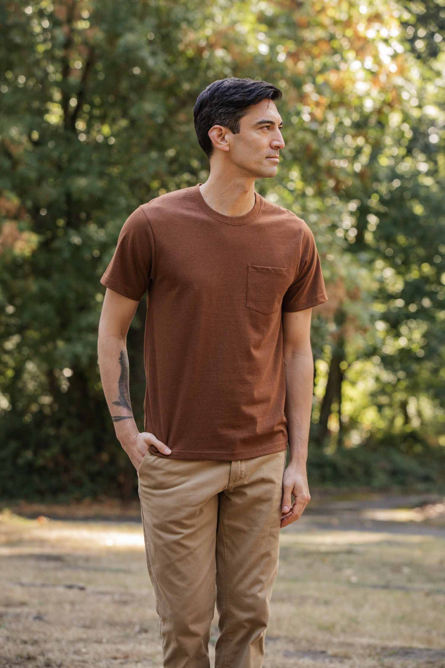 A man stands in a sunlit park wearing the Bridge & Burn Recycled Pocket Tee in Bronze and tan pants. He has short dark hair, a tattoo on his left forearm, and looks to his left with one hand in his pocket. Trees fill the background.