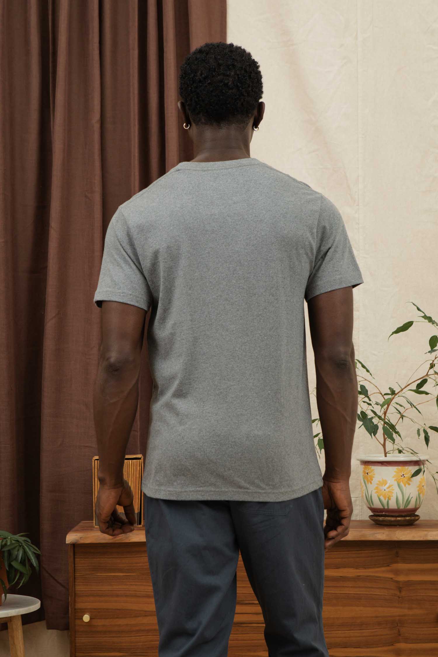 A man with short curly hair faces away, wearing the Bridge & Burn Recycled Pocket Tee in Grey Heather and dark pants, standing by a wooden cabinet with potted plants against brown curtains and a cream wall.