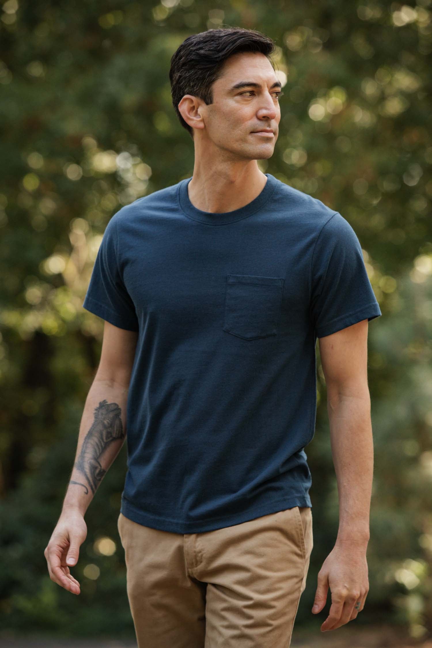 A man with short dark hair and a forearm tattoo stands outdoors among greenery, wearing tan pants and the Bridge & Burn Recycled Pocket Tee in Cobalt, giving the scene a subtle vintage feel as he looks to his left.