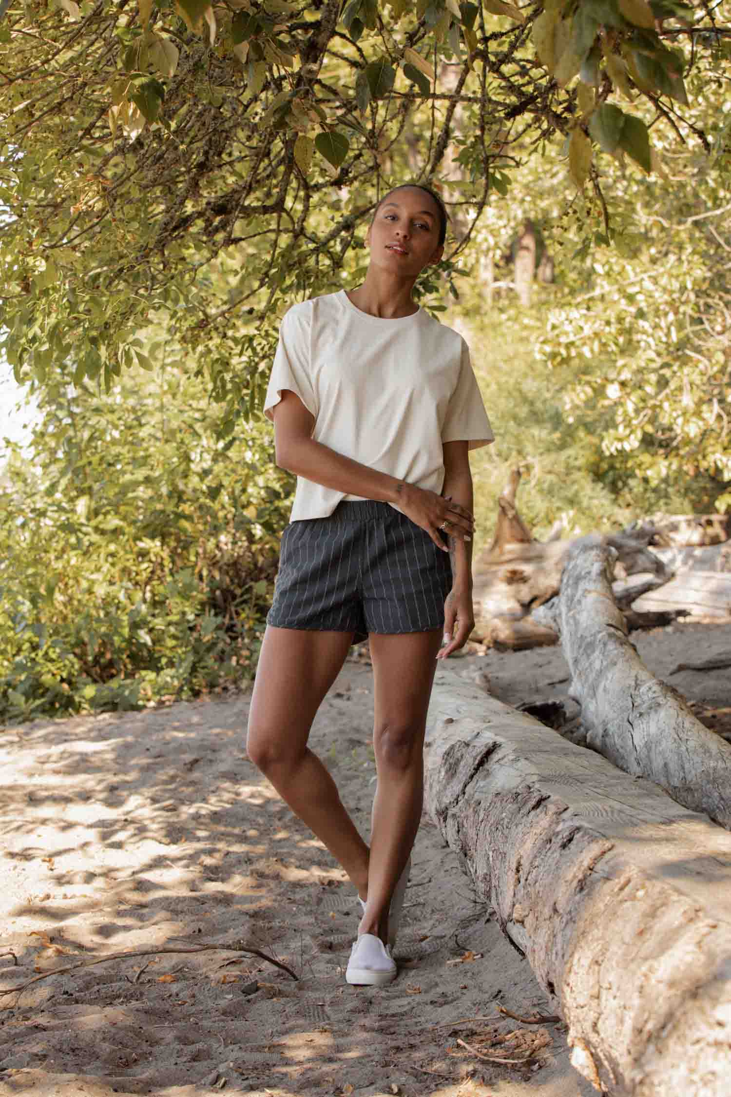 A woman stands on a wooded path beside a fallen tree, wearing the Bridge & Burn Recycled Boxy Tee in Ivory, striped shorts, and white sneakers as sunlight filters through green leaves above her.