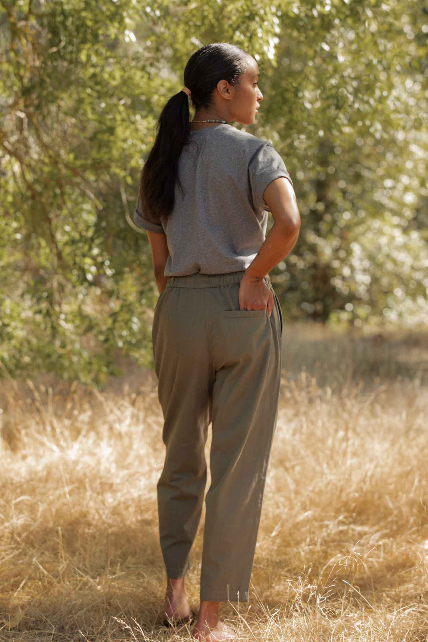 A woman with a ponytail stands outside on dry grass, facing away, wearing the Bridge & Burn Recycled Boxy Tee in Grey Heather and olive-green pants, one hand in her back pocket with trees in the background.
