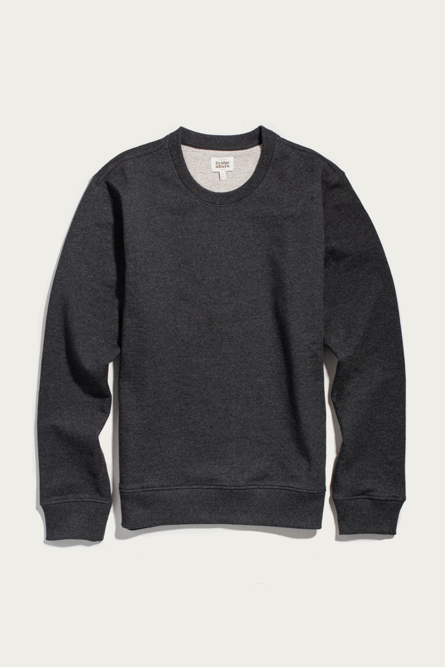 The Hayden Crew Sweatshirt in Graphite by Bridge & Burn is a plain dark gray crew neck sweatshirt with long sleeves and ribbed cuffs, crafted from soft recycled cotton, displayed on a white background.