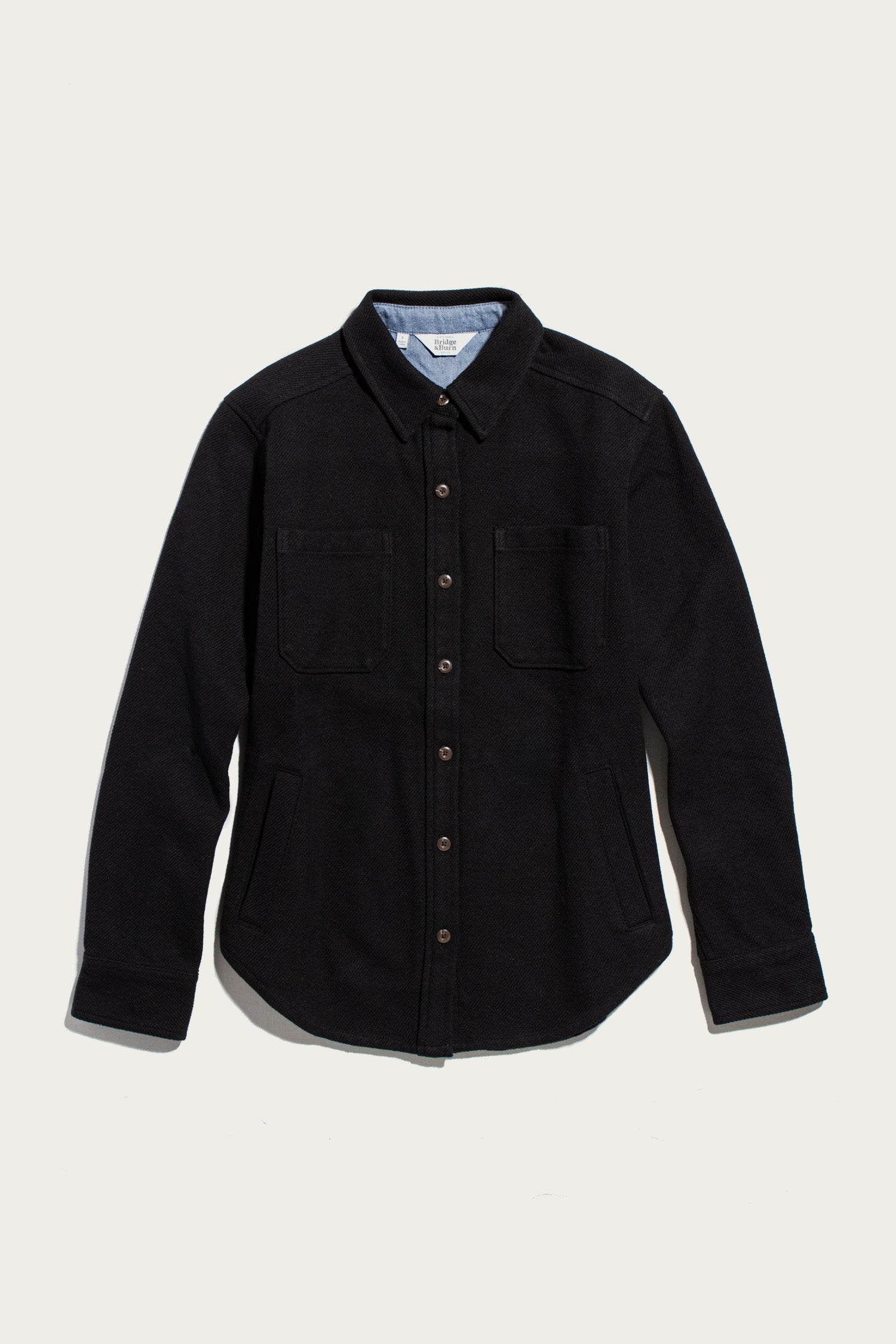 The Dawson Overshirt in Ink Stone by Bridge & Burn is a black button-up shirt jacket crafted from recycled cotton, featuring long sleeves, a collar, two chest pockets, and convenient side pockets. Shown against a plain white background, this modern recycled overshirt is perfect for those who value sustainable clothing.