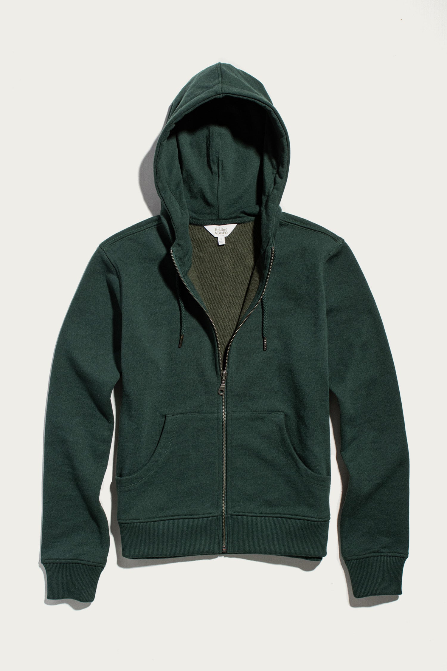 Bridge & Burn’s Mason Hoodie in Juniper is a dark green, zip-up sustainable hoodie featuring a hood, front pockets, and long sleeves, crafted from recycled French terry sweatshirt material and displayed flat on a white background.