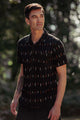 A man with short dark hair stands outdoors in the Bridge & Burn Harbor Slim Shirt in Dotted Horizon, a black summerweight cotton shirt with a geometric pattern, showing off his tattooed forearm against blurred greenery.