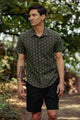 A man with short dark hair and an arm tattoo stands among lush greenery, wearing the Bridge & Burn Harbor Slim Shirt in Thyme Arrows and black shorts. The tailored fit shirt features a subtle pattern, perfect for summer.