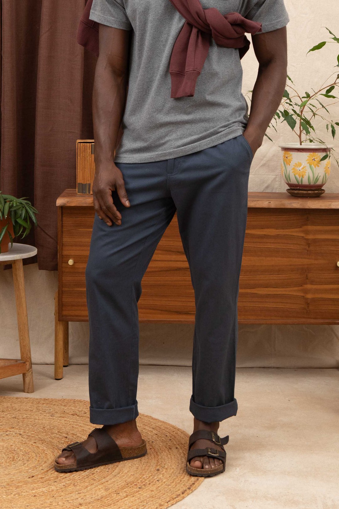 A person stands indoors wearing a grey t-shirt, maroon sweatshirt over their shoulders, Bridge & Burn Hayes Pant in Dark Slate, and black sandals on a woven rug near a wooden sideboard with plants.