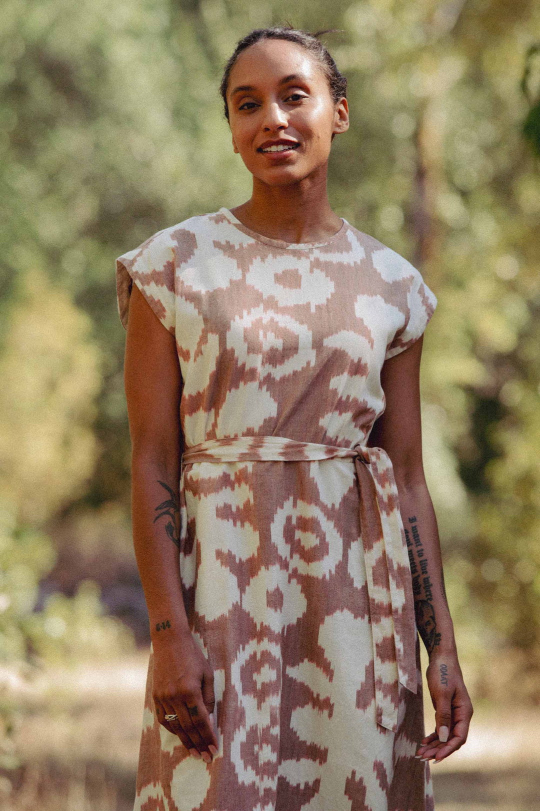 Outdoors, a person wears the Bridge & Burn Eloise Dress in Pop Leaf—a beige and white patterned 100% cotton dress with short sleeves and a waist tie—smiling slightly with arm tattoos, against a backdrop of blurred greenery.