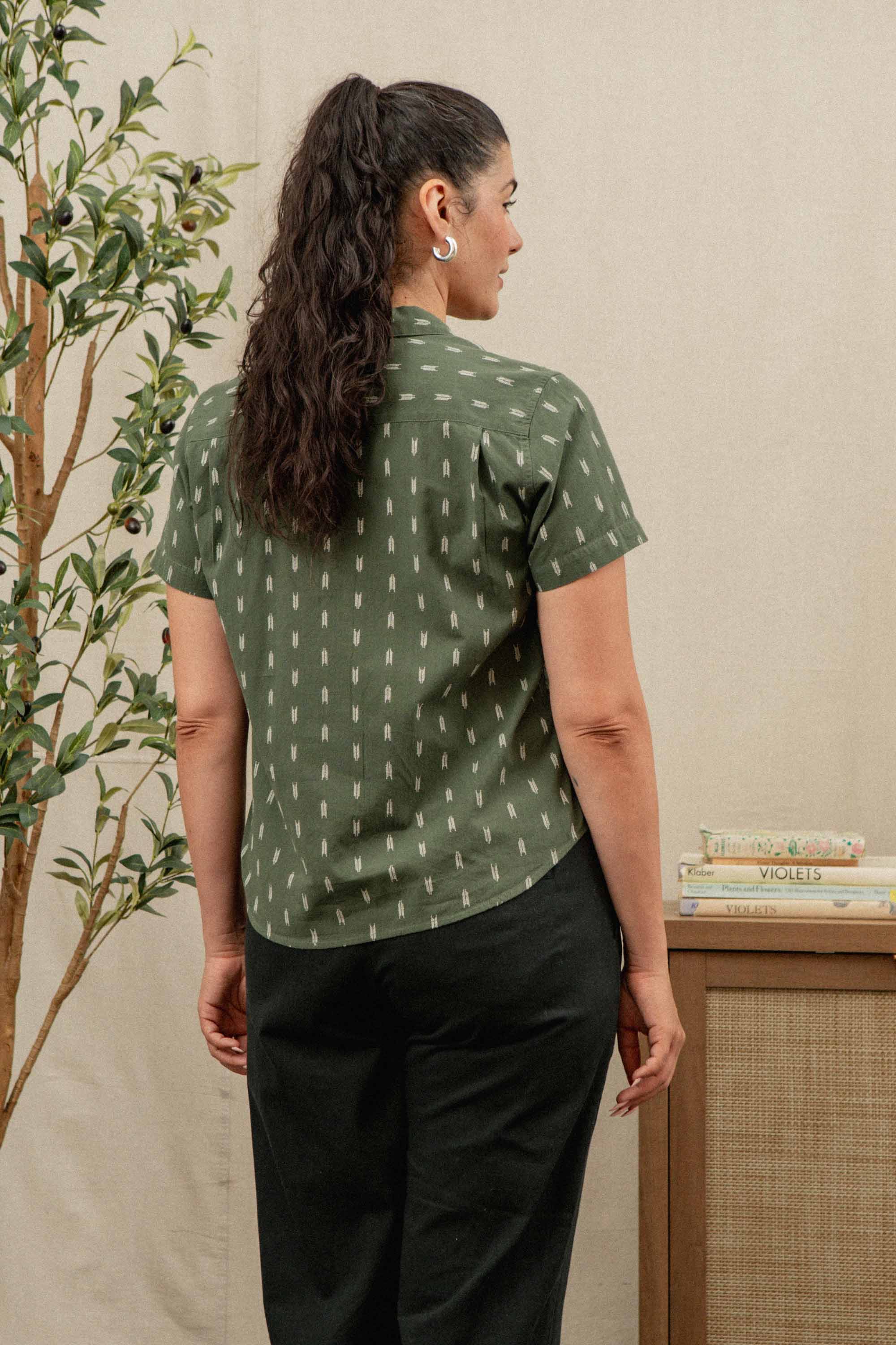 A woman with long dark hair in a ponytail stands with her back to the camera, wearing Bridge & Burn's Lark Pant in Soft Black and a green patterned shirt. A small table with books and a potted plant sits beside her.