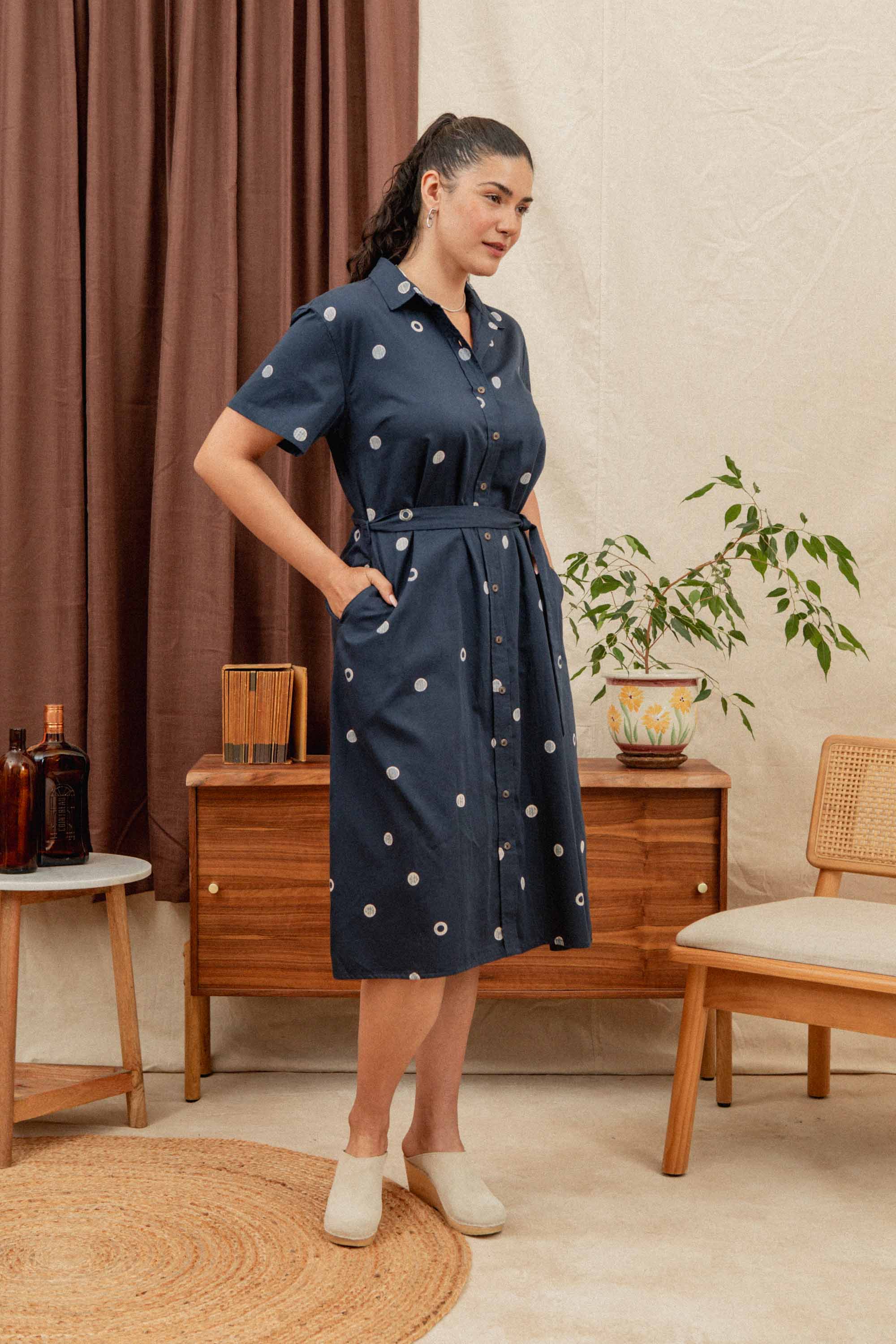 The Sylvie Dress / Lunar Polka by Bridge & Burn is a navy blue, oversized organic cotton shirtdress with short sleeves, a collar, a matching waist tie, and white circular patterns. It is shown laid flat on a plain background.