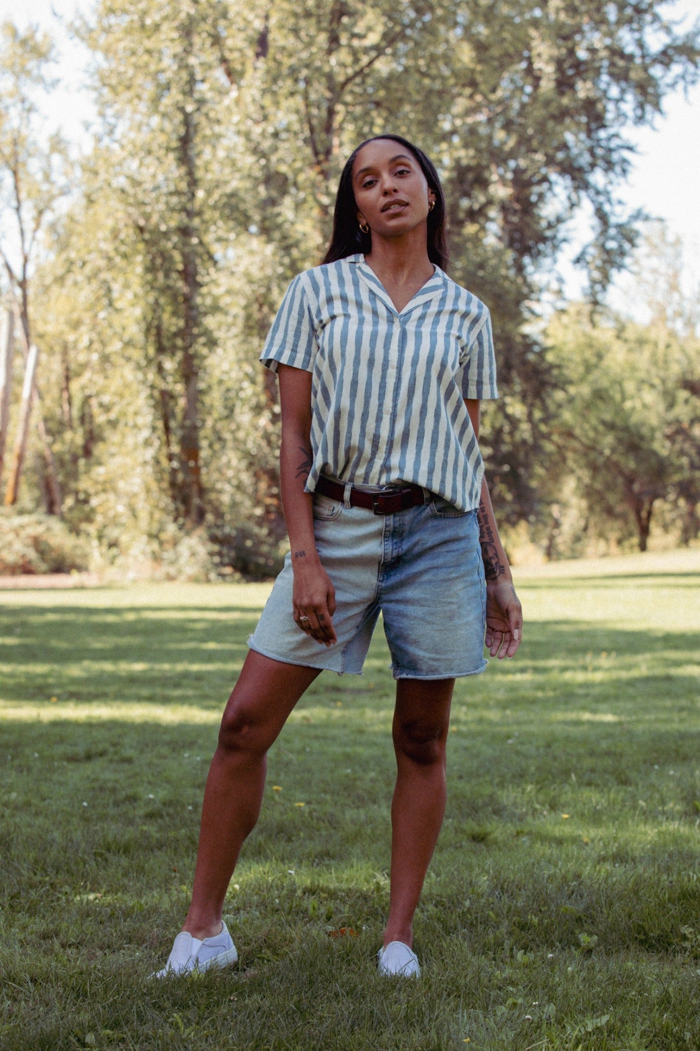 A woman stands on grass in a sunlit park, wearing the Bridge & Burn Wren Shirt in Beach Stripe, styled with denim shorts and white shoes. Trees and greenery fill the background on a clear day.
