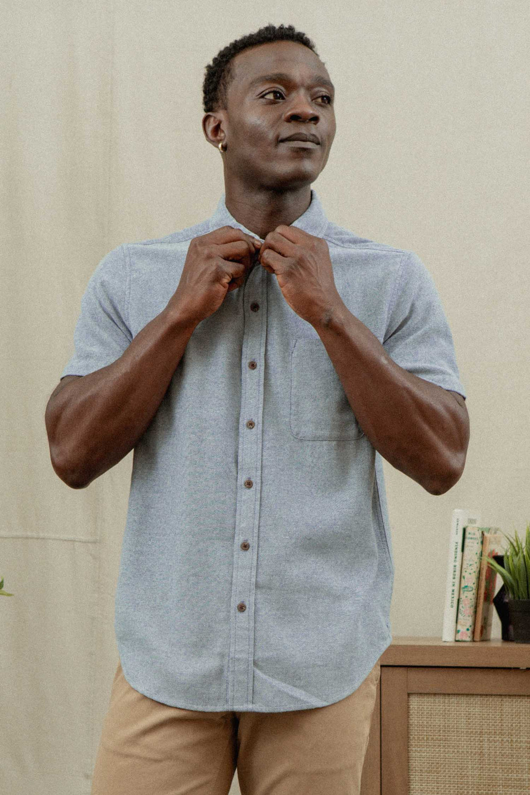 A man stands indoors, buttoning the collar of his Bridge & Burn Bryce Shirt in Denim Twill, paired with tan pants. Behind him are a cabinet with books and a small plant, set against a neutral background.