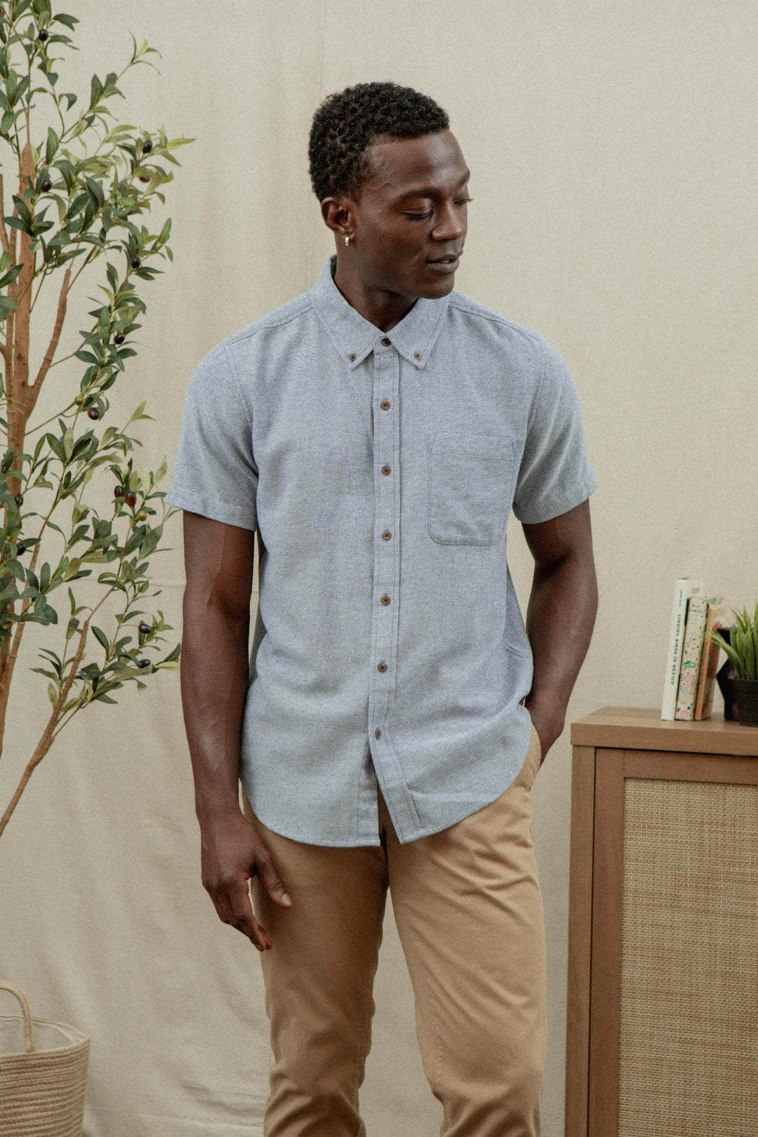A man wearing the Bridge & Burn Bryce Shirt in Denim Twill and tan pants stands indoors near a plant and cabinet, looking down with one hand in his pocket.