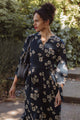 A woman with curly hair in a bun wears the Bridge & Burn Fiona Dress in Night Garden, a navy cream floral dress, paired with gold earrings and a black shoulder bag as she walks outdoors on a sunny day surrounded by greenery.