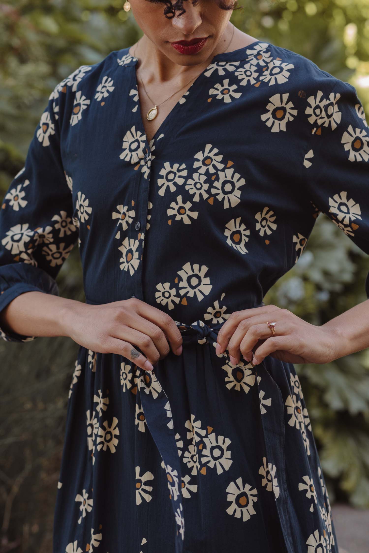 Wearing the Bridge & Burn Fiona Dress / Night Garden—a navy blue gathered sleeve dress with a white floral pattern—a person with curly hair and red lipstick adjusts the waist tie while standing outdoors among lush greenery.