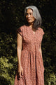 A person with long, wavy gray hair stands outdoors in lush greenery, wearing the Bridge & Burn Ira Dress in Desert Rain—an organic cotton, rust-orange patterned dress with short sleeves.