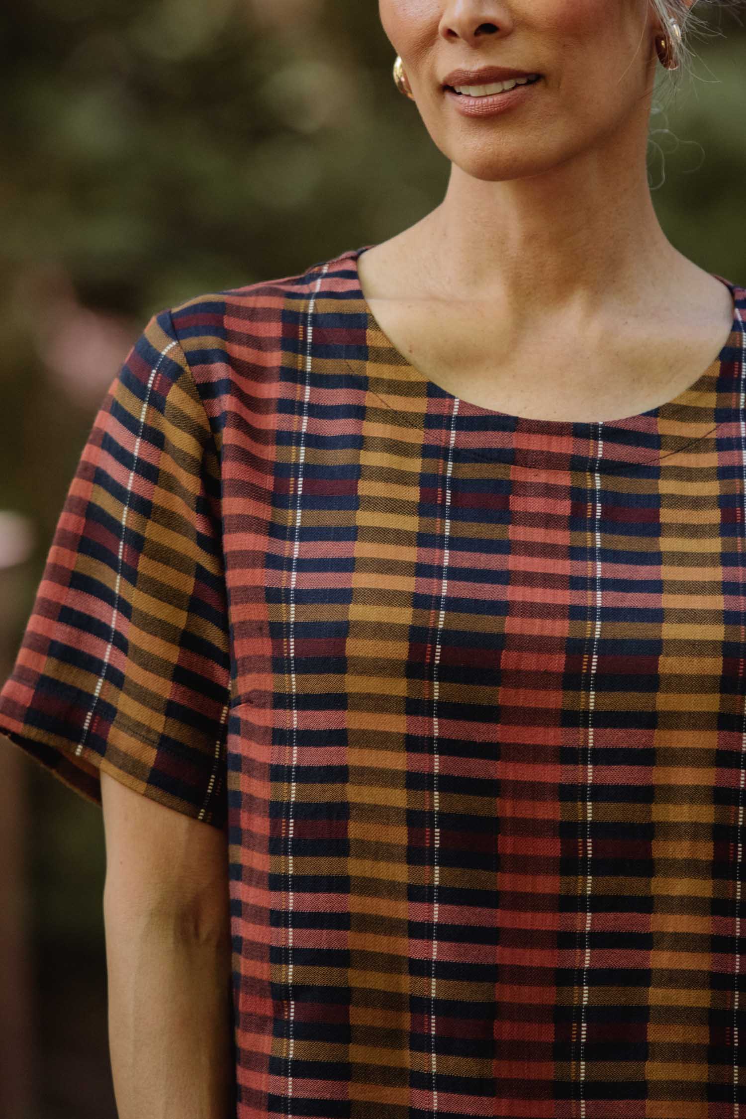 A woman wears the Bridge & Burn Leona Shirt in Alpenglow Stripe, a boxy blouse with short sleeves and a cropped hem, featuring multicolored stripes. Made from eco-friendly viscose, she stands outdoors with only her upper body visible.