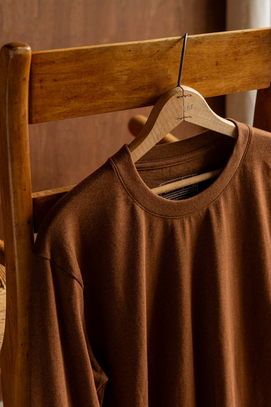 The Barney Long Sleeve Tee in Bronze by Bridge & Burn—a relaxed fit crew neck crafted from recycled cotton—is draped over the back of a wooden chair on a wooden hanger, with warm light accentuating the rich textures of both the fabric and wood.