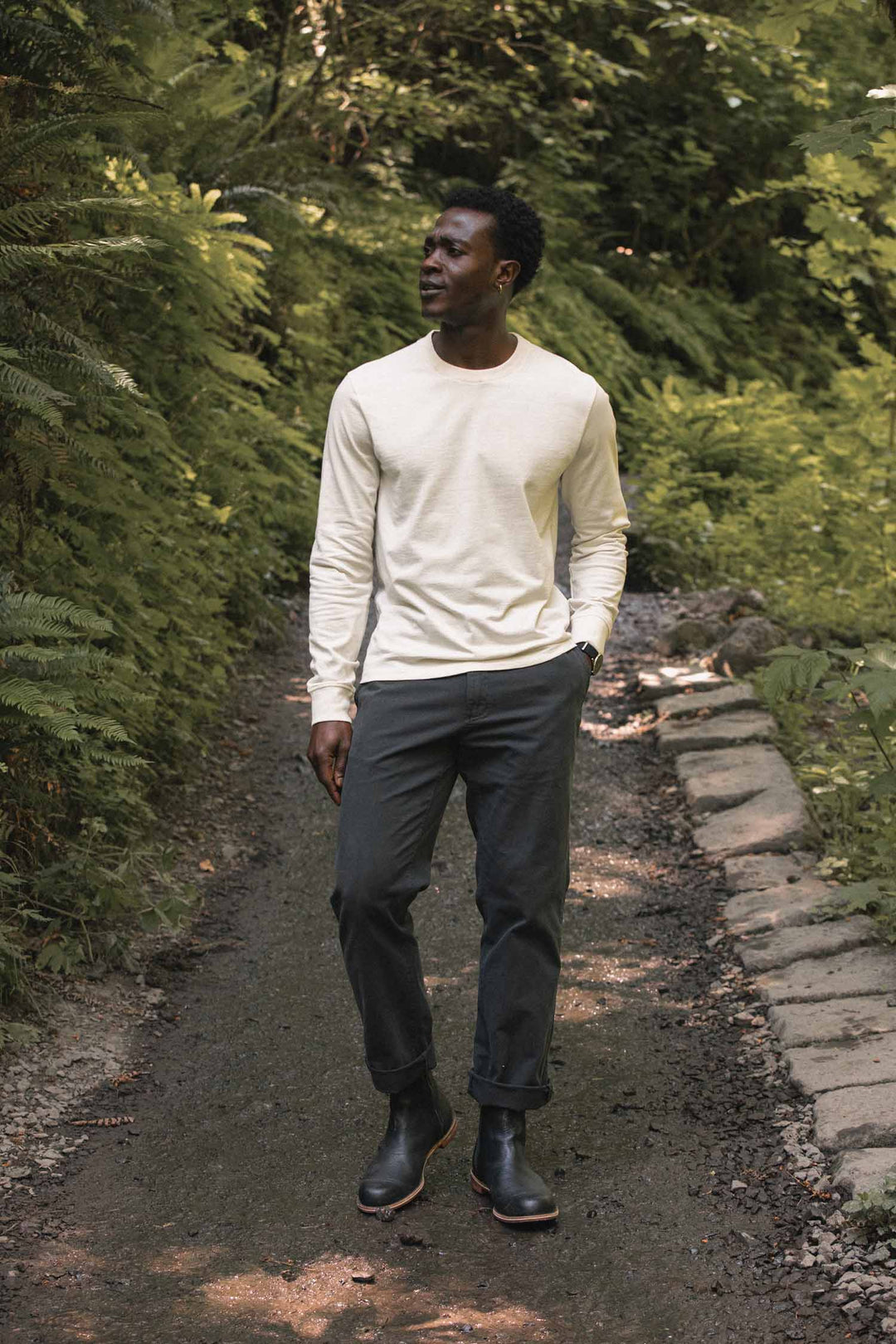 Wearing the Bridge & Burn Barney Long Sleeve Tee in Ivory, a man in relaxed-fit dark pants strolls along a shaded forest path lined with green ferns and trees, one hand tucked in his pocket.