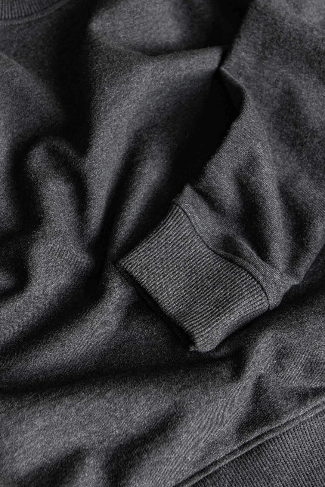 A close-up of the Bridge & Burn Hayden Crew Sweatshirt in Graphite highlights its ribbed cuff and soft recycled cotton fabric texture, with natural folds and shadows adding depth to the material.