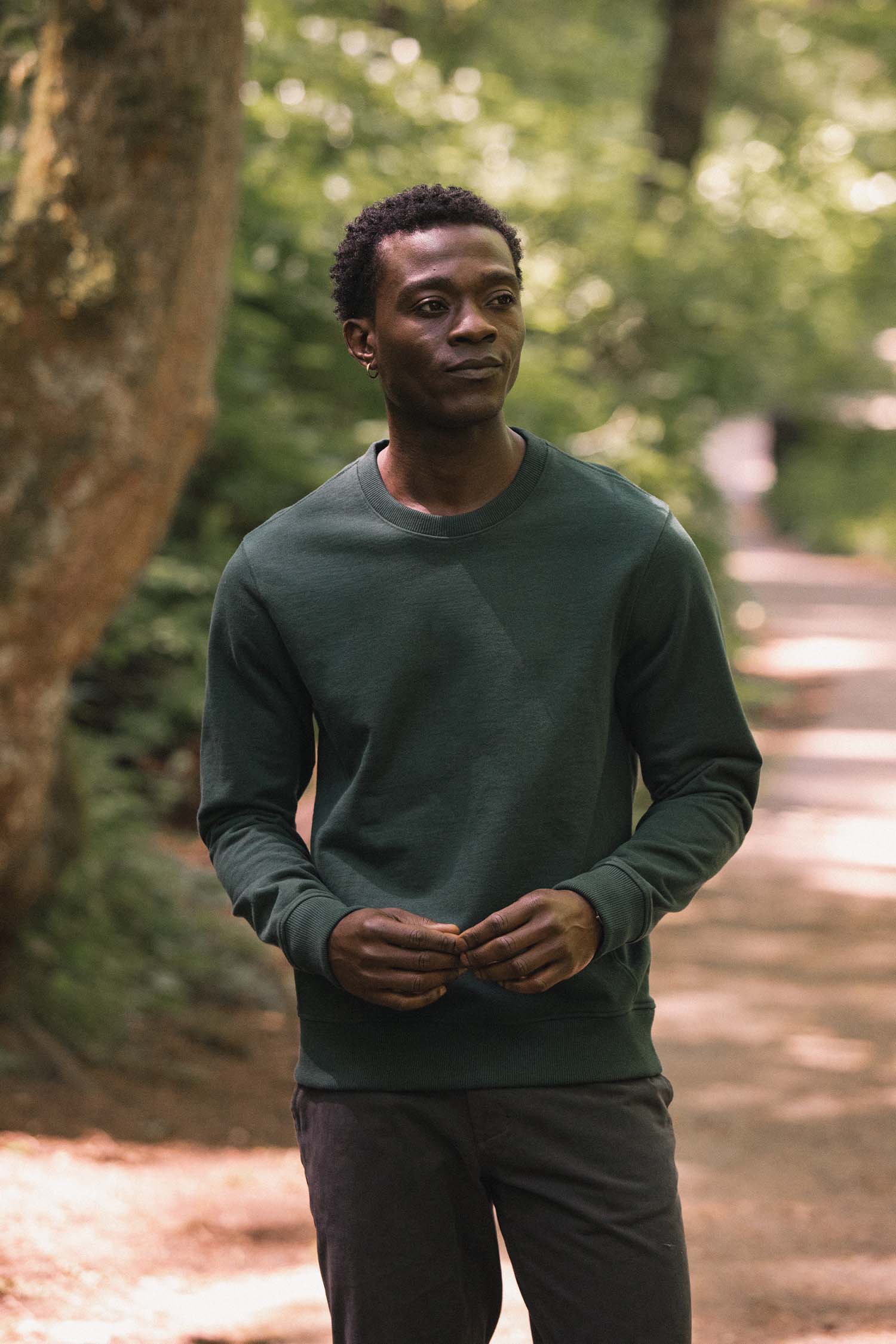 A man wearing the Bridge & Burn Hayden Crew Sweatshirt in Juniper stands on a forest path, holding something in his hands as sunlight filters through the trees behind him.