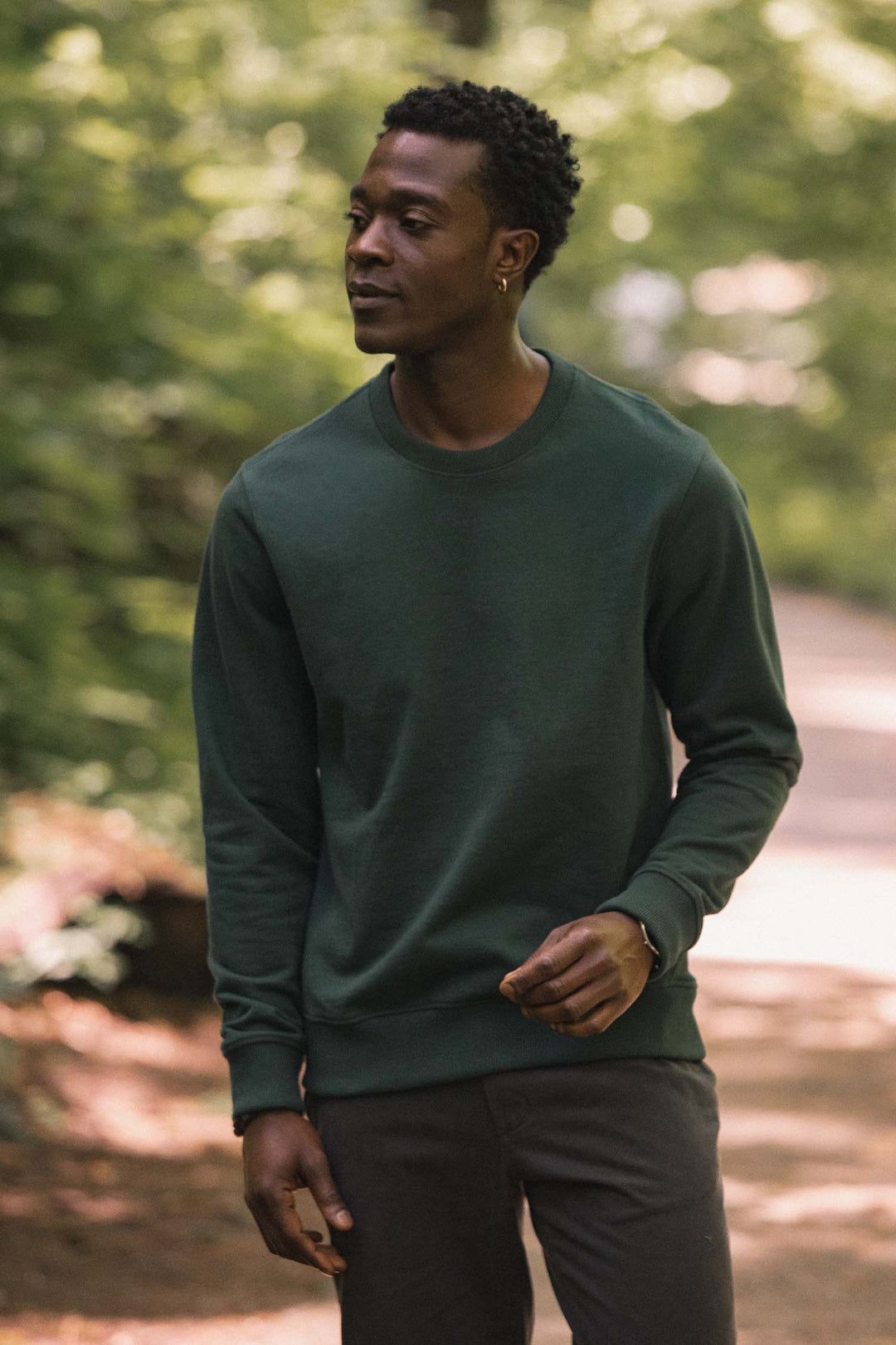 A man wearing the Bridge & Burn Hayden Crew Sweatshirt in Juniper and gray pants stands outdoors on a sunlit path surrounded by green trees and foliage.