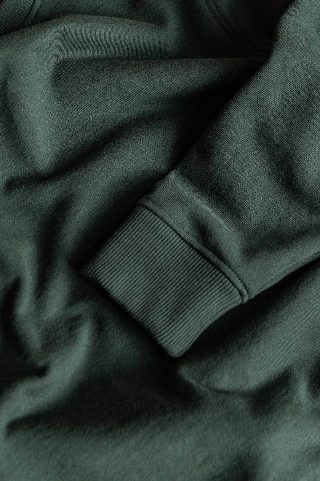 Close-up of the Hayden Crew Sweatshirt in Juniper by Bridge & Burn, featuring a ribbed cuff on a forest green recycled cotton sleeve, displayed against a matching fabric background.