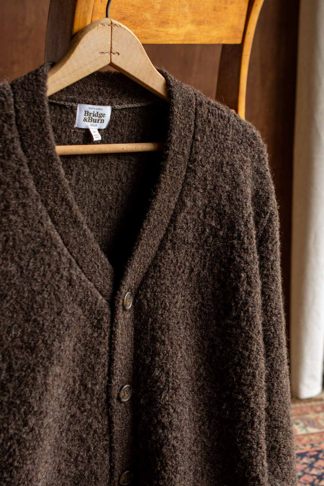 A textured knit dark olive Mateo Cardigan by Bridge & Burn, featuring large buttons and patch pockets, hangs on a wooden hanger draped over the back of a wooden chair. The Bridge & Burn label is visible inside the collar.