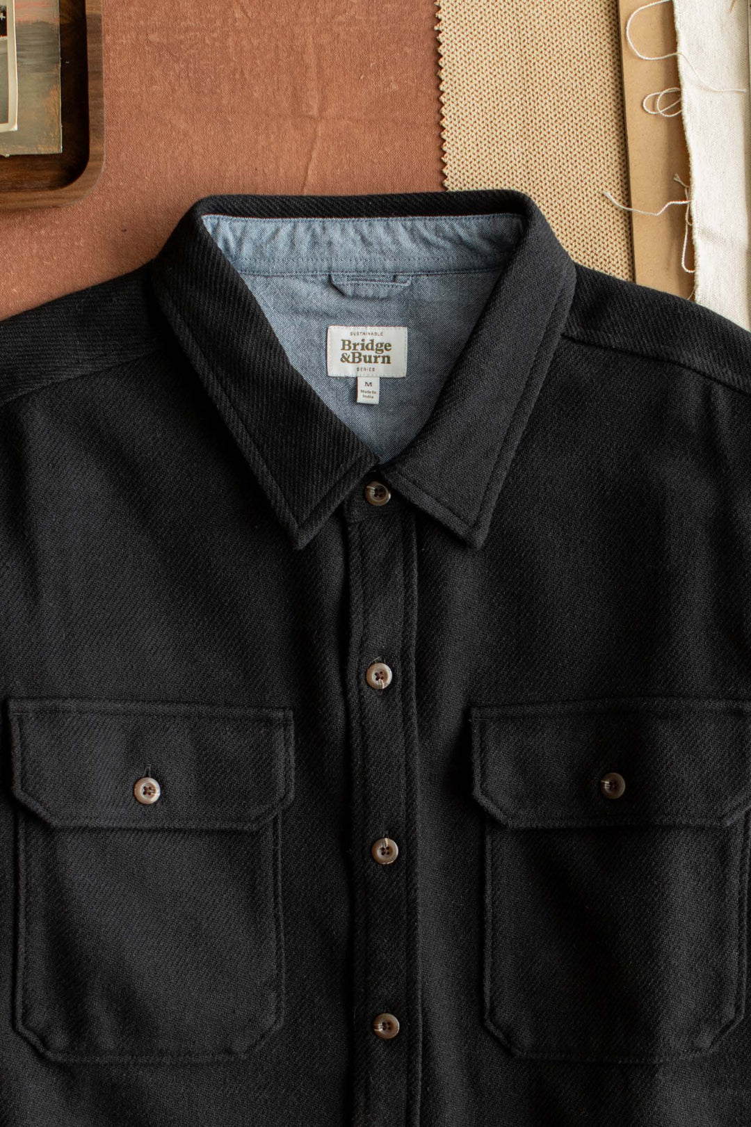 The Pointer Overshirt in Ink Stone by Bridge & Burn—a black twill overshirt with front chest pockets—is laid flat on a brown surface, accompanied by tan fabric swatches and white thread, exemplifying an elevated staple for those who value sustainable clothing.