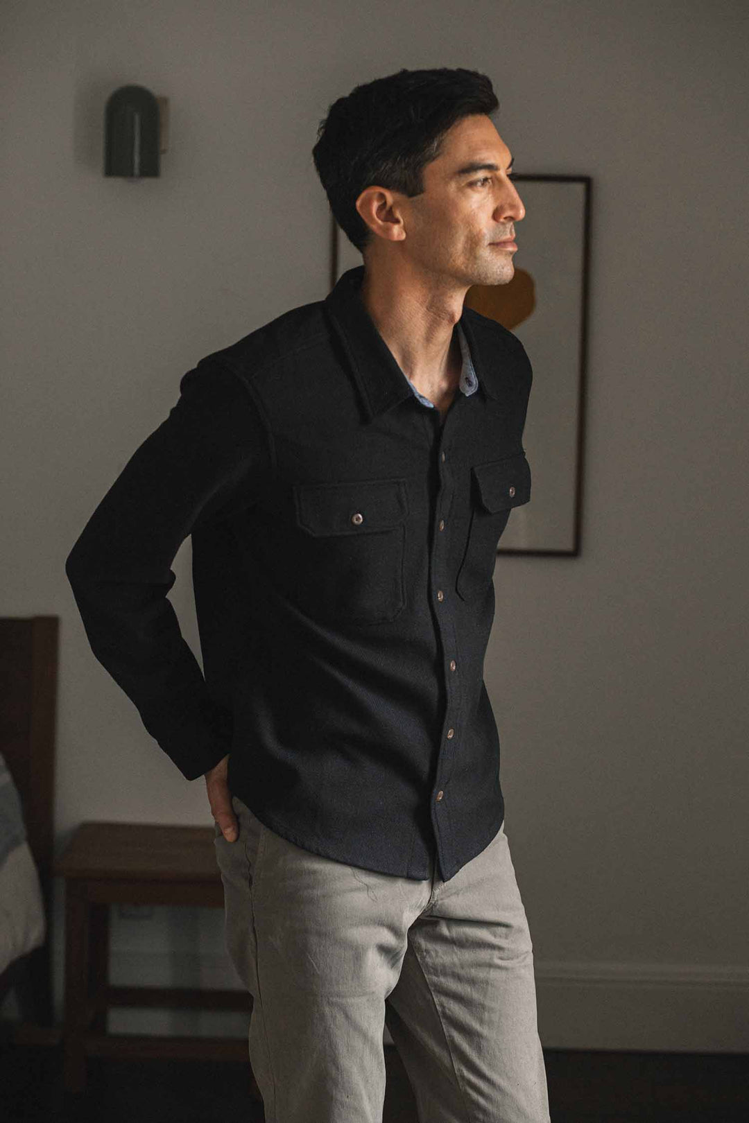 A man wearing the Bridge & Burn Pointer Overshirt in Ink Stone and light gray pants stands indoors, looking to the side with his hands in his pockets. The background includes a bed, a chair, and a framed picture on the wall.