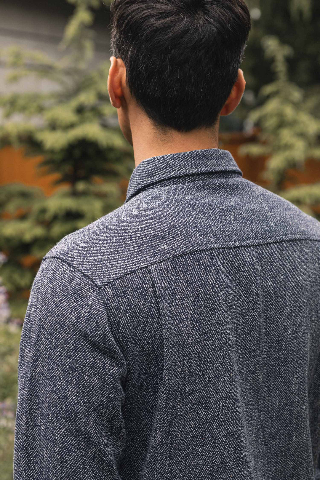 A person with short dark hair is seen from behind, wearing the Bridge & Burn Pointer Overshirt in Ocean Marl, crafted from recycled fibers. Blurred trees and a wooden fence are visible in the background.
