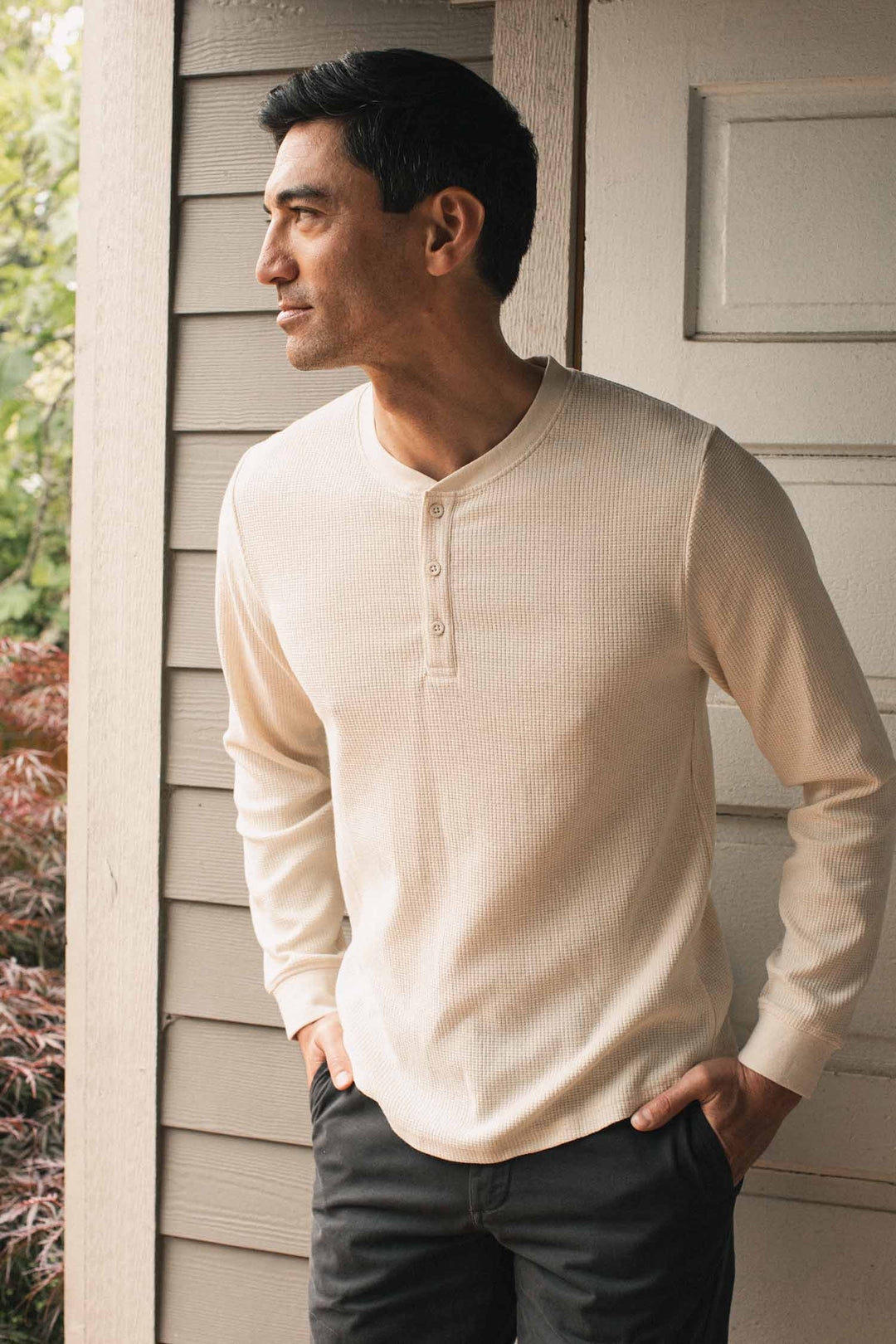 A man wearing the Bridge & Burn Men’s Recycled Waffle Henley in Ivory stands in front of a light wooden wall, looking to his left with one hand in his pocket.
