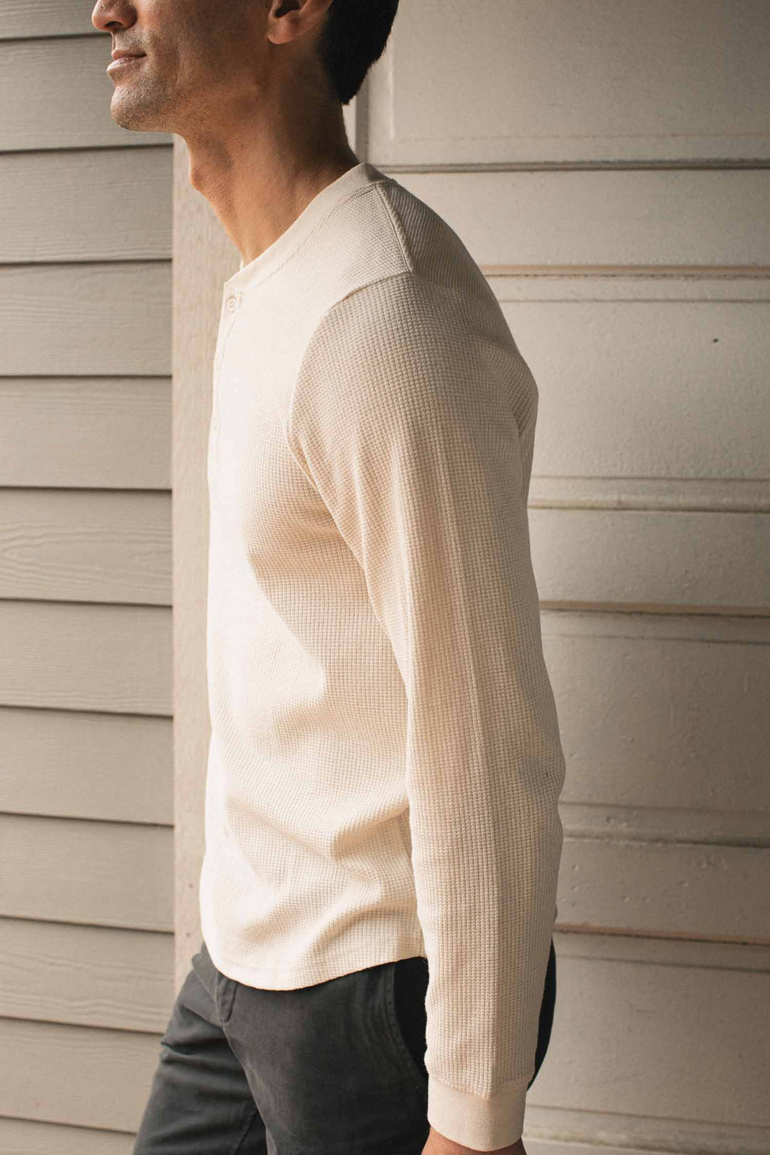 A man models the Bridge & Burn Men’s Recycled Waffle Henley in Ivory, standing sideways near a light wood-paneled wall with part of his face out of frame.