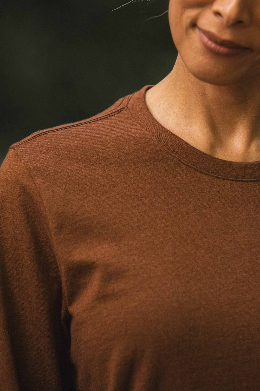 A person wearing the Bridge & Burn Audrey Long Sleeve Tee in Bronze, shown from the shoulders up with a partial view of their smiling face. The background is dark and blurred.