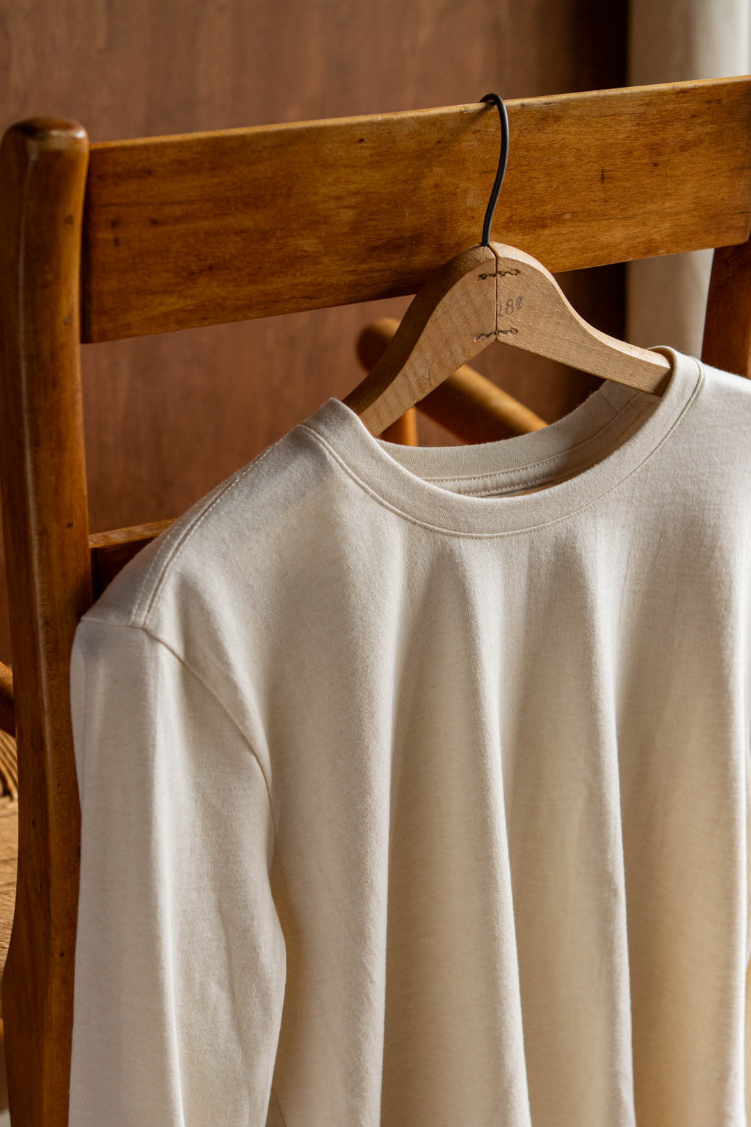 An ivory Audrey Long Sleeve Tee from Bridge & Burn, crafted from recycled cotton, hangs on a wooden hanger draped over the back of a wooden chair against a warm brown background, highlighting its effortless and relaxed fit.