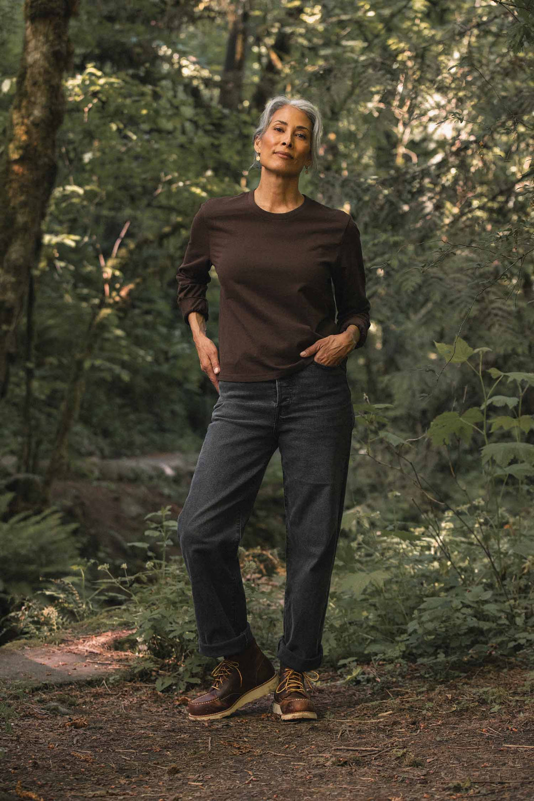 A person with short gray hair stands on a forest path, wearing a relaxed-fit Audrey Long Sleeve Tee in Truffle by Bridge & Burn, paired with dark jeans and brown boots. Sunlight filters through the trees, creating a peaceful, natural setting.