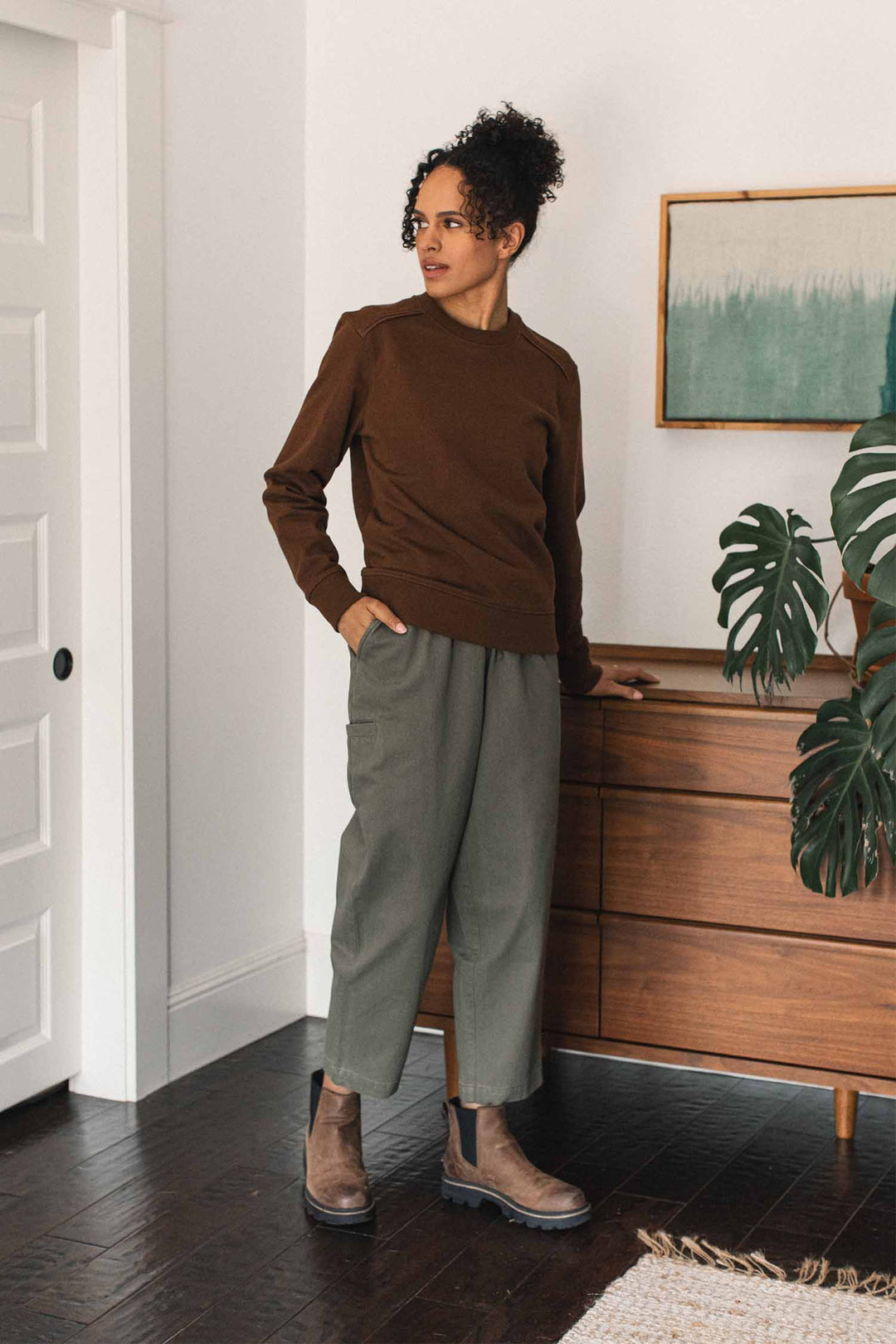 A person stands indoors beside a wooden dresser, wearing the Bridge & Burn Bailey Crew Sweatshirt in Cumin, paired with green pants and brown boots. A green plant and framed art sit atop the dresser, creating a modern and minimalistic setting.