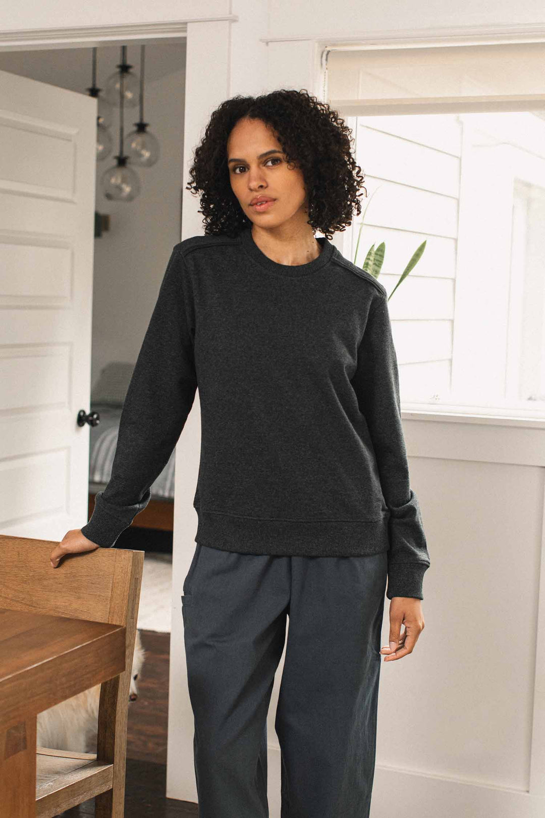 A person with short curly hair stands indoors, resting one hand on a wooden table, wearing the Bridge & Burn Bailey Crew Sweatshirt in Graphite and pants, with a white door and windows in the background.