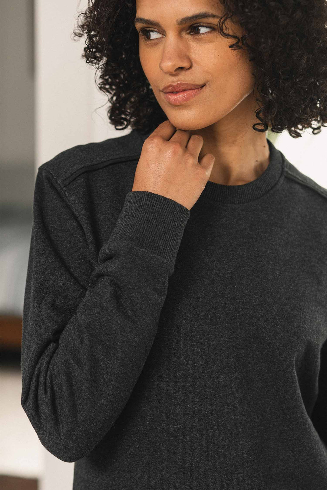 A woman with curly hair, wearing the Bridge & Burn Bailey Crew Sweatshirt in Graphite, looks to the side with a slight smile, gently touching her neck against a softly blurred background.