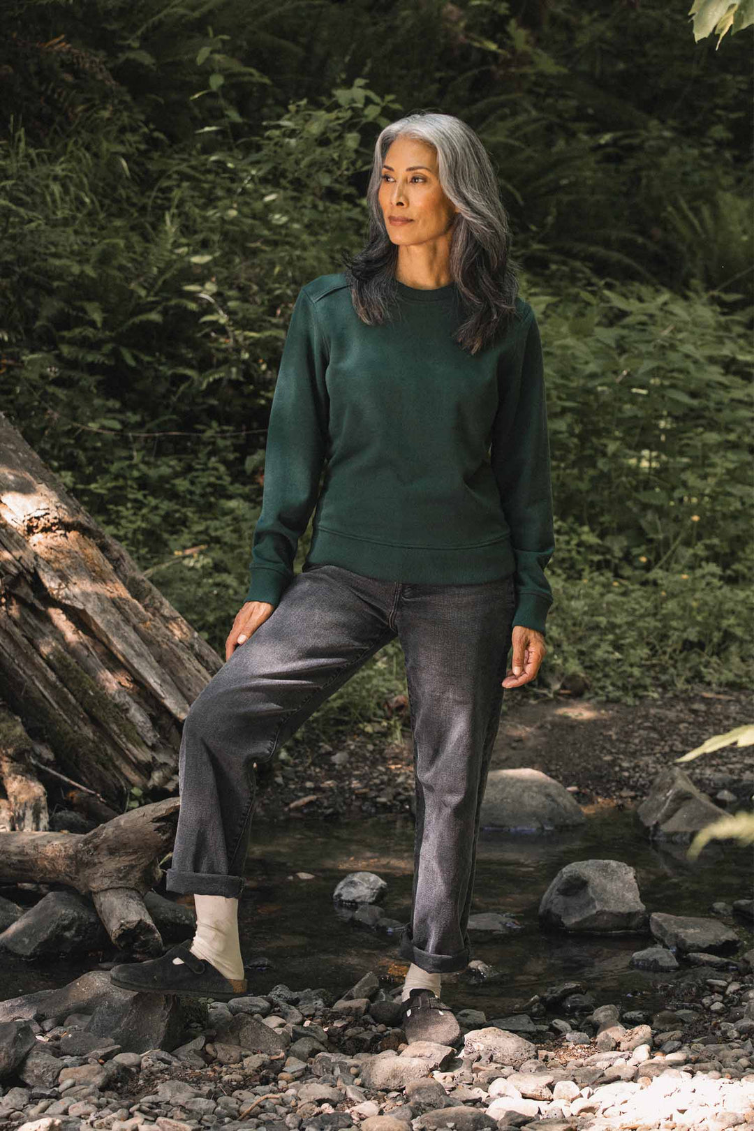 A woman with long gray hair stands on rocks in a forest, wearing the Bridge & Burn Bailey Crew Sweatshirt in Juniper and dark jeans, one foot wrapped in a white bandage or sock, surrounded by greenery and sunlight.